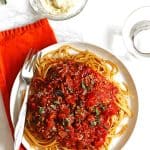 Spaghetti sauce and noodles on a white plate with fresh herbs garnishing it. A bowl of grated parmesan cheese is next to it with a spoon.