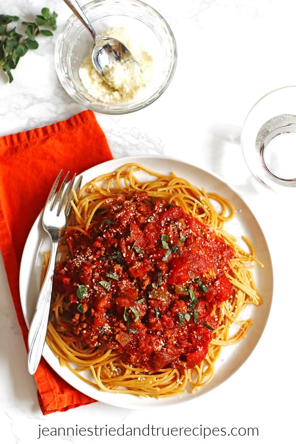 Spaghetti sauce and noodles on a white plate with fresh herbs garnishing it. A bowl of grated parmesan cheese is next to it with a spoon.