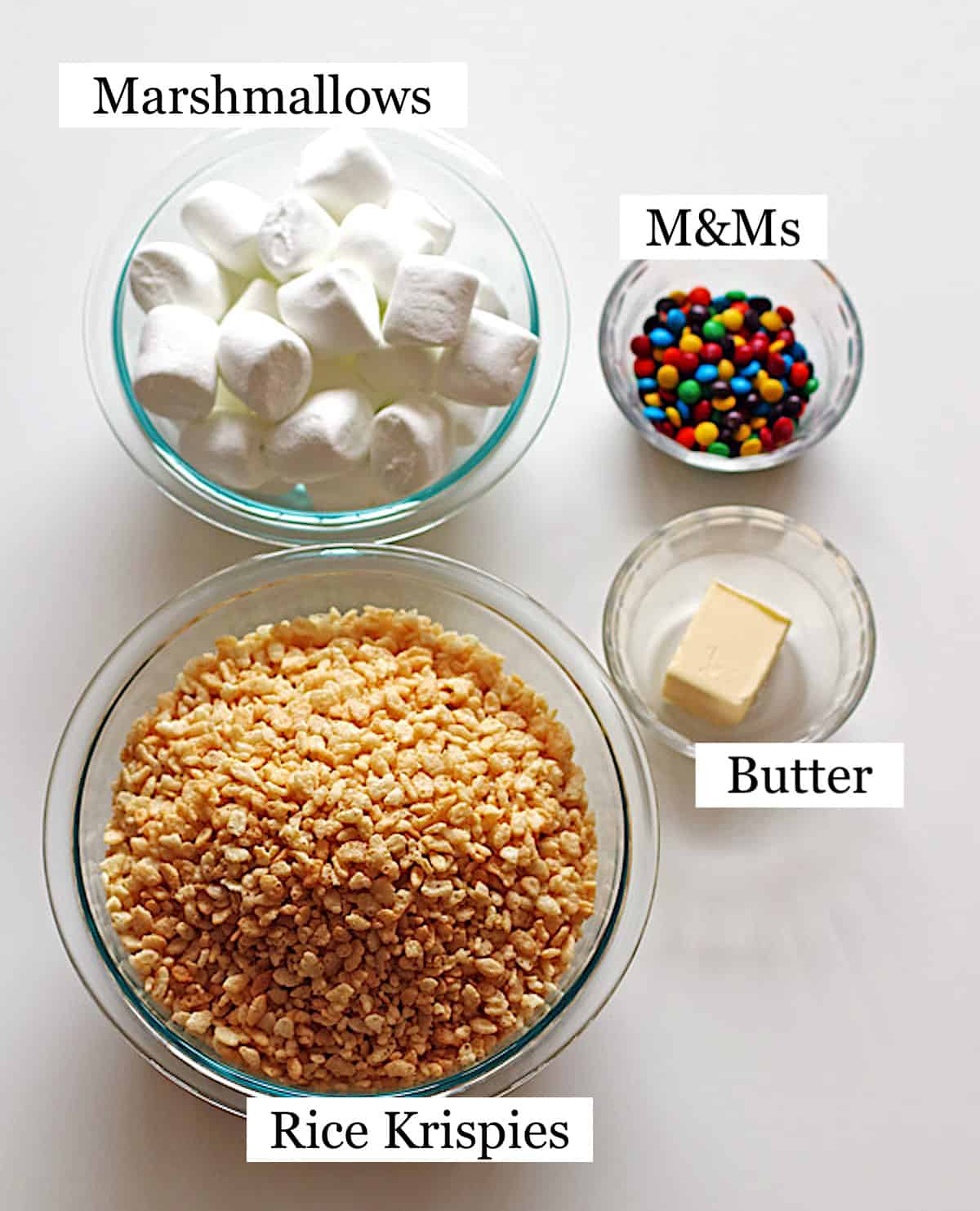 Rice Krispie cereal, marshmallows, M&M candy and butter in four separate clear bowls