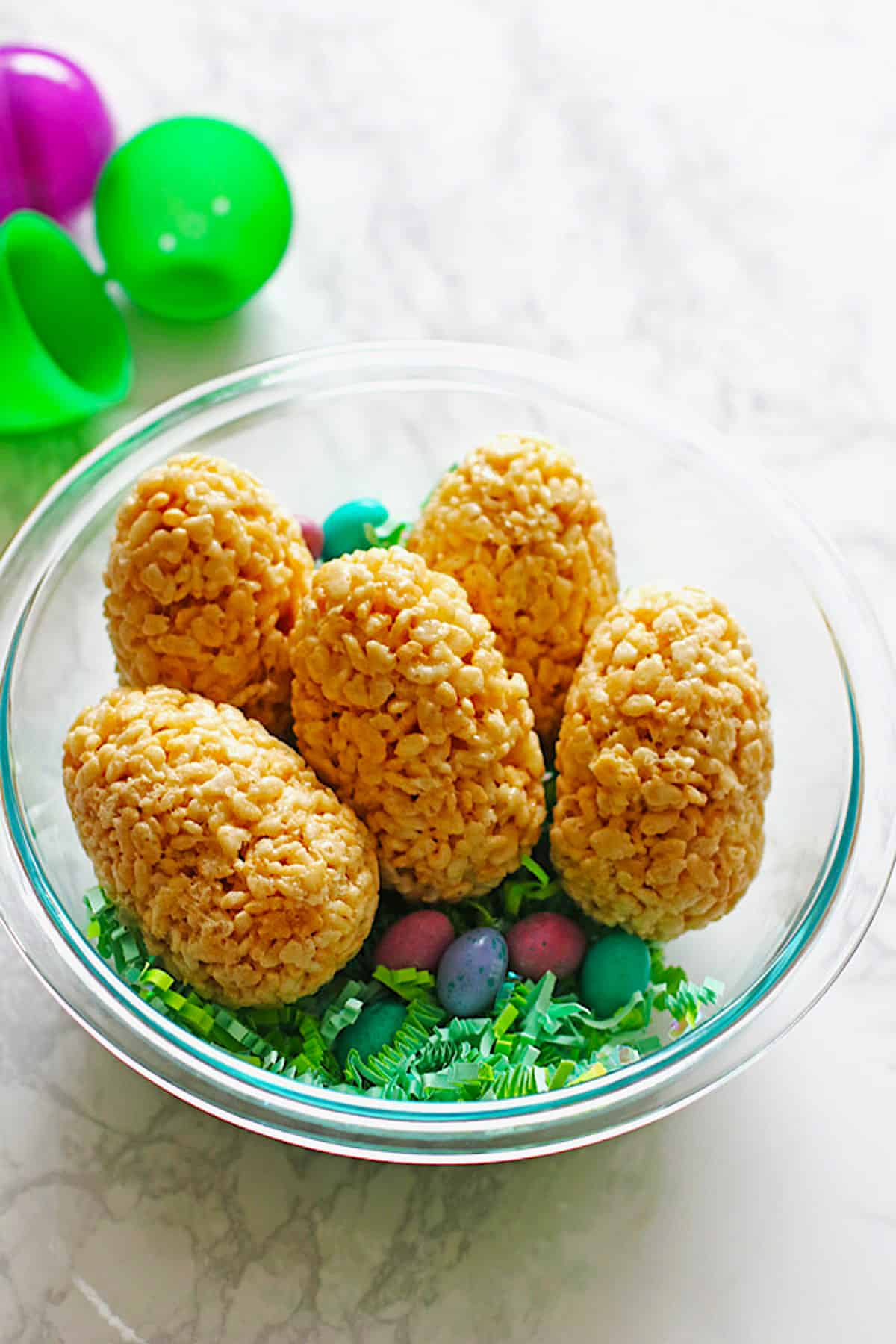 Easter eggs made out of rice krispie treats placed in a clear bowl with green Easter grass and candy