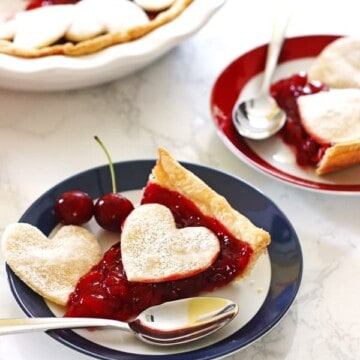 Cherry Pie slices on red and blue rimmed plates with spoons. Pie in dish next to the plates.