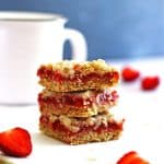 Bars made of strawberries and rhubarb cut into squares. There are three bars stacked on each other next to a white mug.