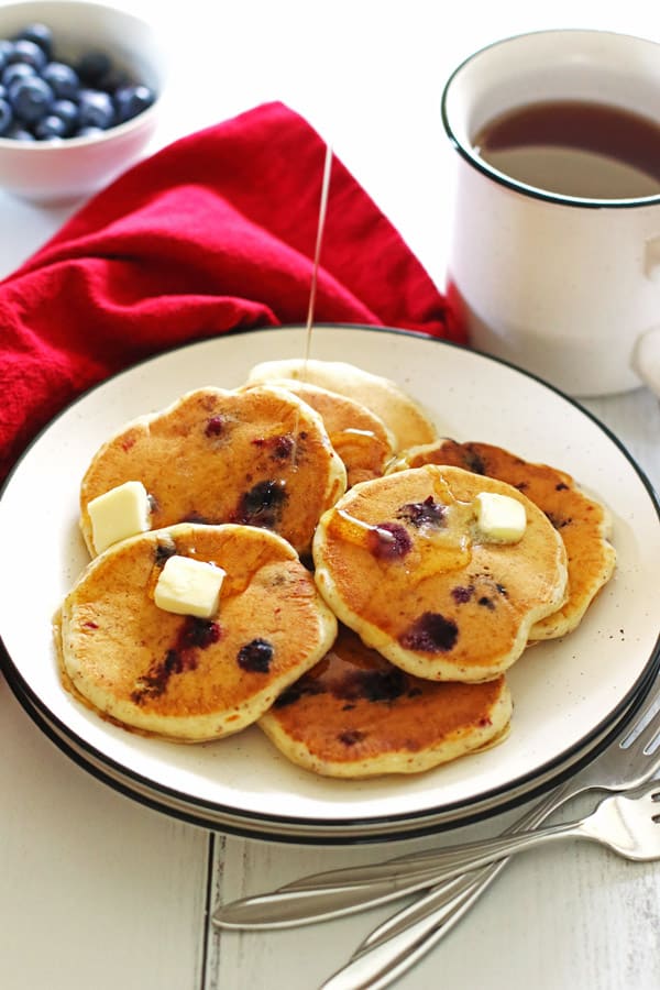 Pancakes made with blueberries on a white plate with sliced butter on top of them and syrup being poured over them