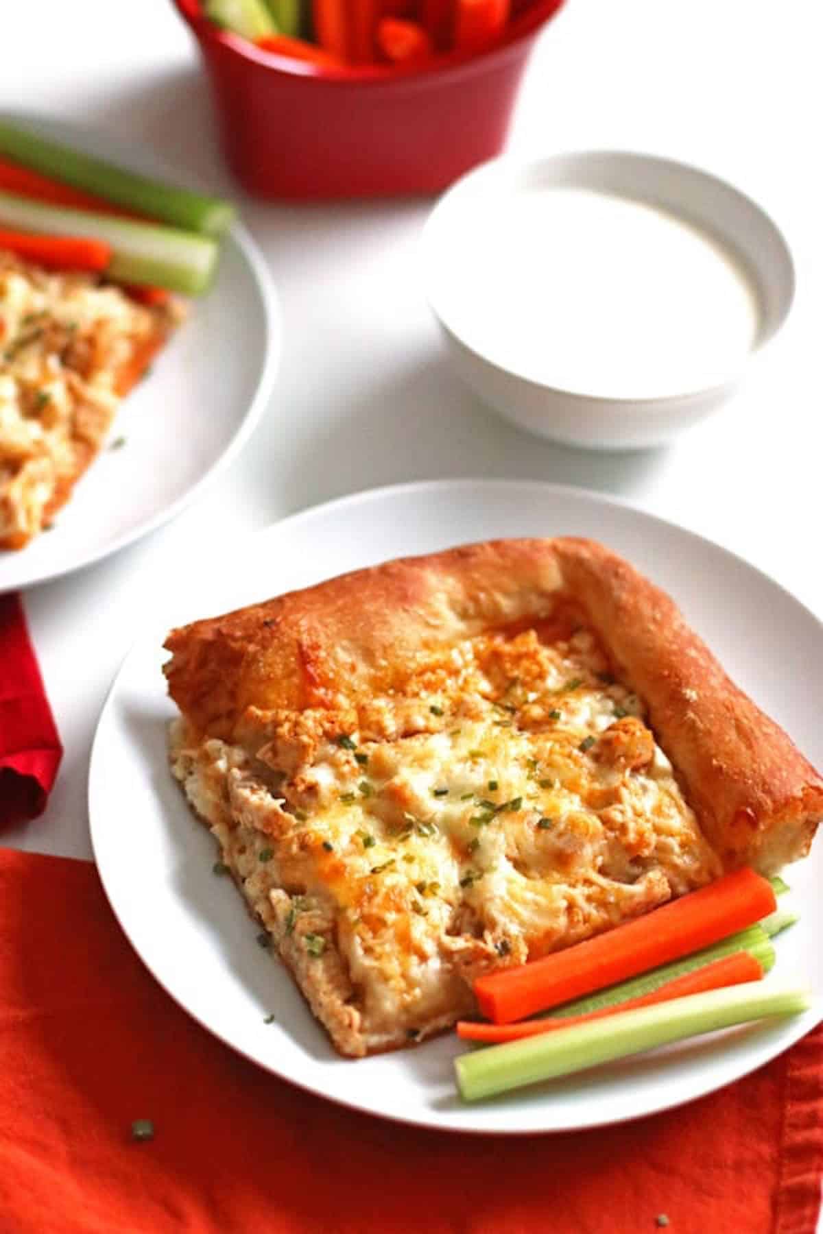 A corner piece of buffalo chicken pizza on a white plate with celery and carrot sticks.