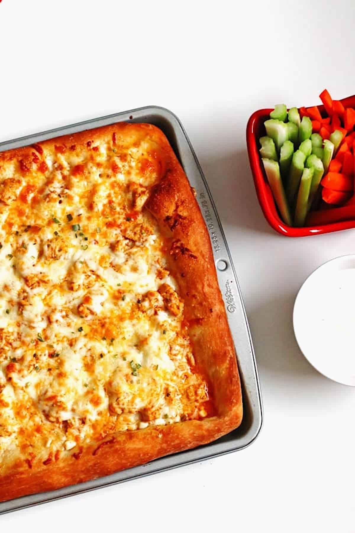 Buffalo Chicken Pizza in baking pan with carrots, celery and blue cheese dressing on the side.