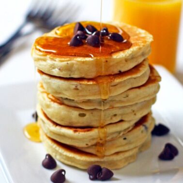 A stack of chocolate chip pancakes with maple syrup on a white plate with a glass of orange juice in the background.