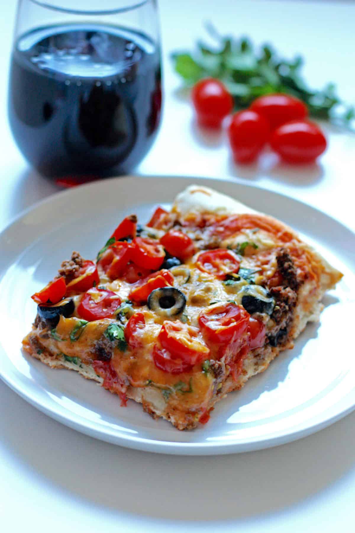 A square slice of taco pizza topped with cheese, black olives, and tomatoes, with a glass of wine behind it.