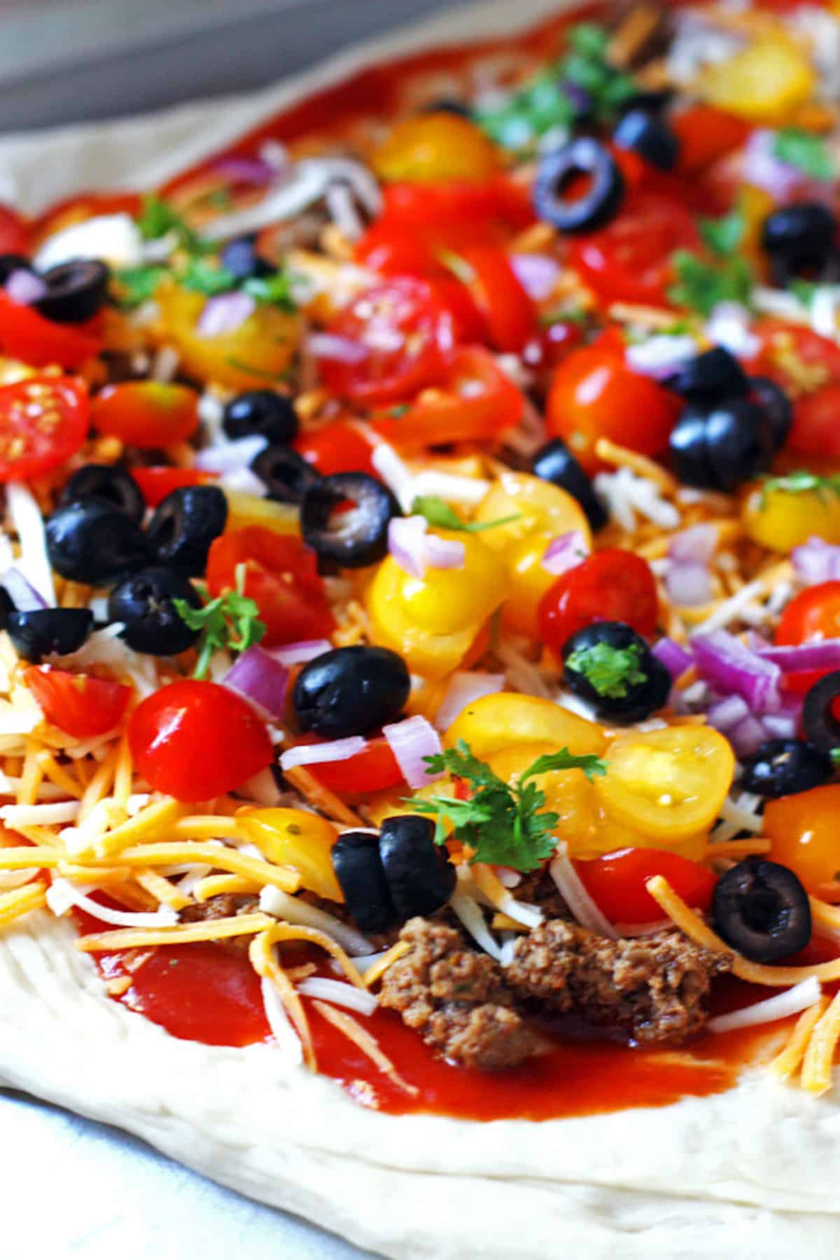 An unbaked taco pizza topped with cheese, tomatoes, ground beef, red onions, and black olives.