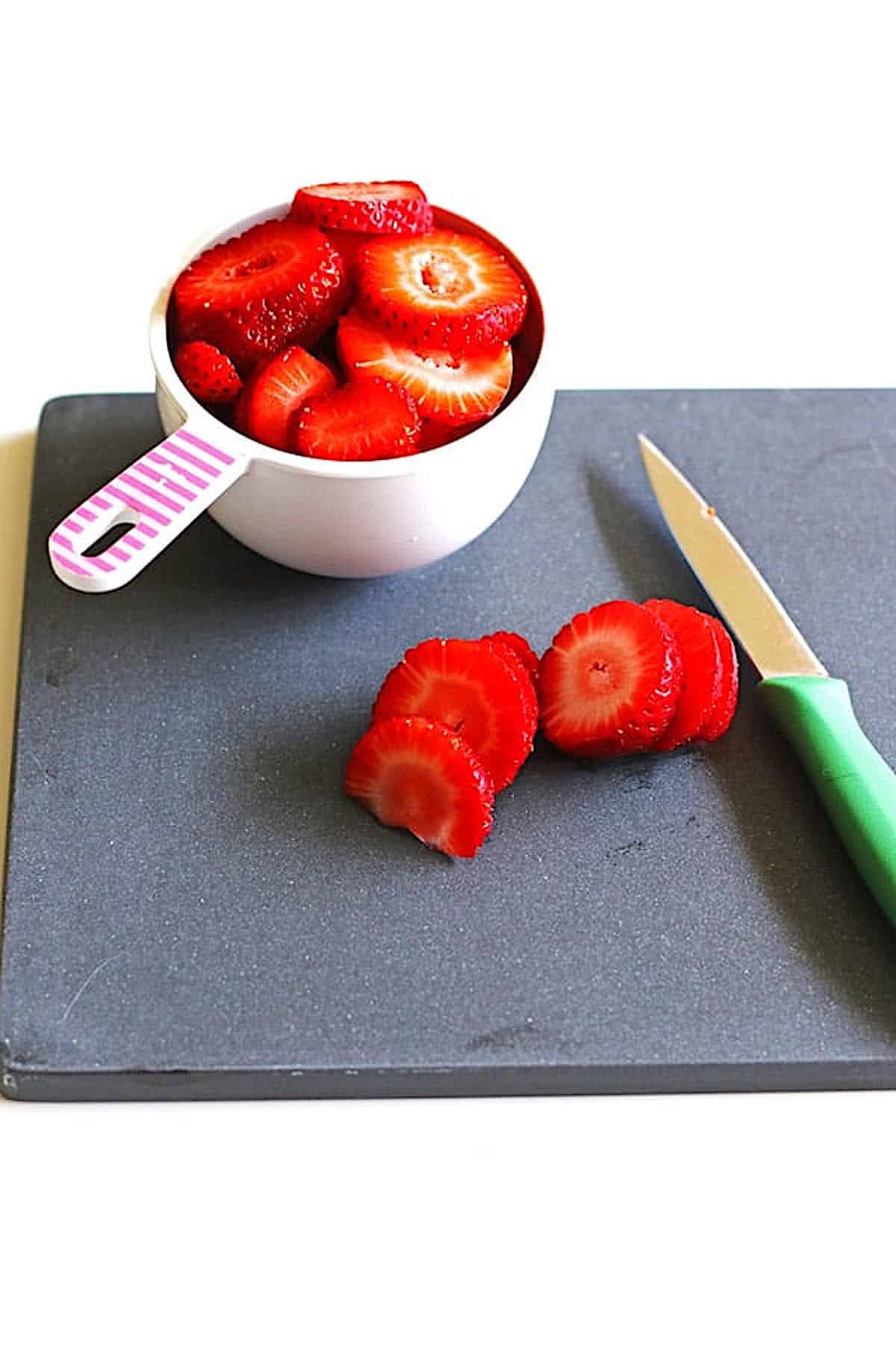 Sliced strawberries in a measuring cup sitting on a black cutting board with a knife