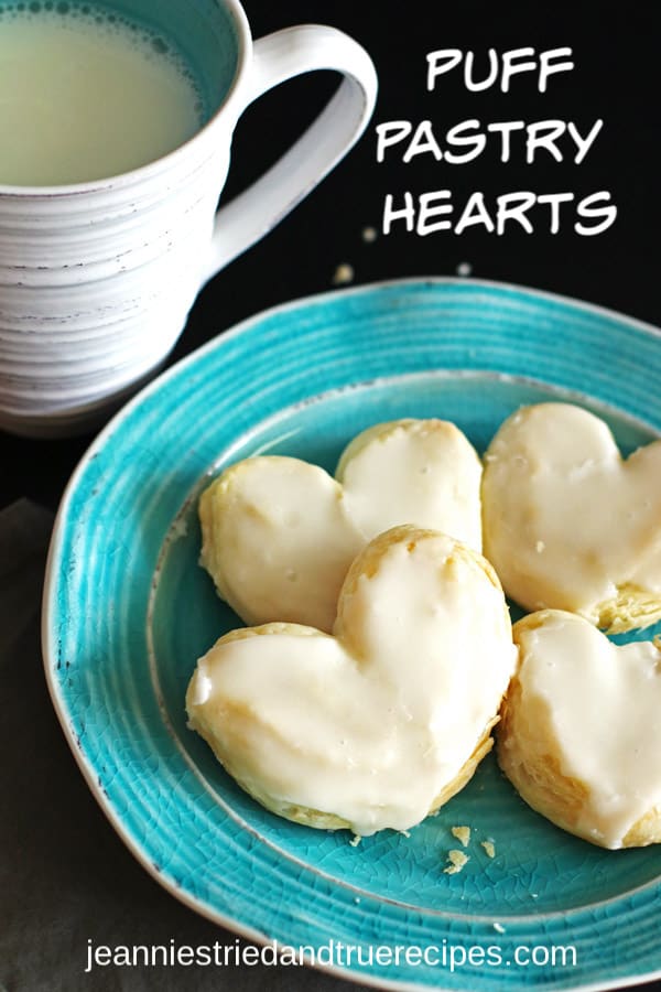 Four Puff pastry in the shape of hearts on a light blue plate with a mug of milk next to it