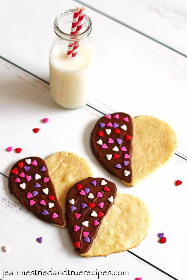 Sugar Cookies made in the shape of hearts that are half dipped in chocolate. They have red, white and pink heart sprinkles on them and are sitting on a table with a glass of milk.