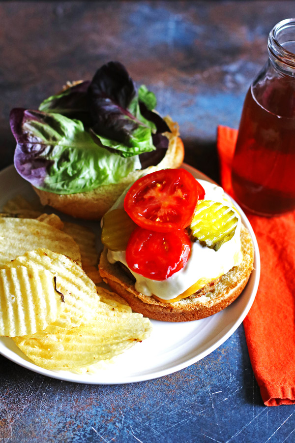 Blue Cheese Burger on a roll on a white plate with chips and pickles on the side.