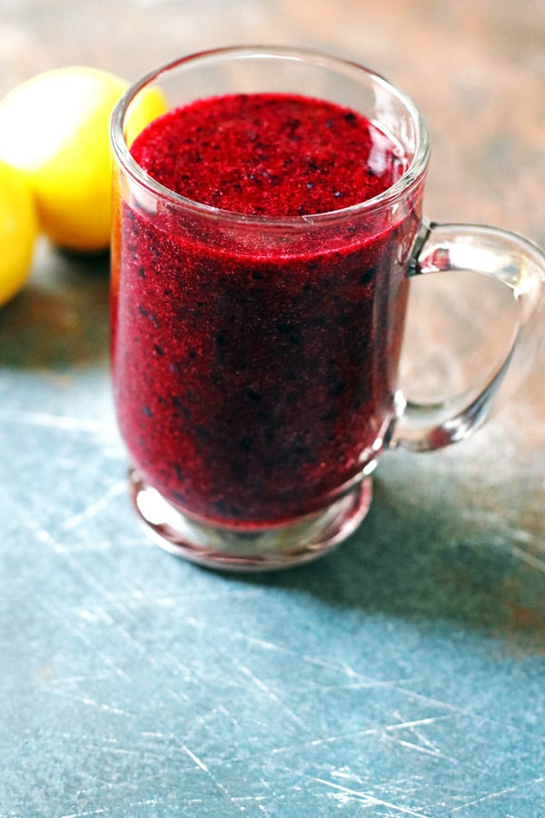 Smoothie made of blueberries and lemon in a clear mug. It is sitting on a table with lemons next to it.