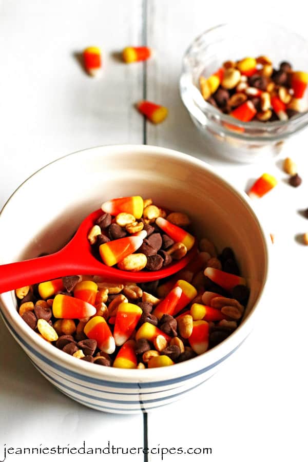 Candy Corn Mix in a white bowl and a large red spoon is being used to serve it. There are small clear pyrex dishes close by with some of the mix in one of them.
