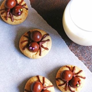 Peanut butter cookies made with candy spiders on top placed next to a glass of milk on a table
