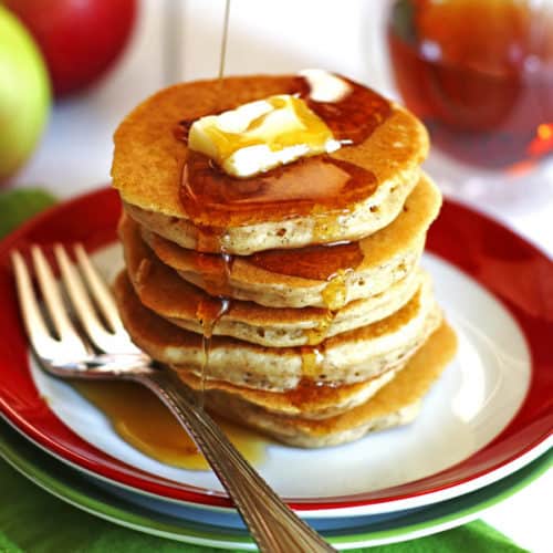 A stack of Apple Pancakes on a plate. There is sliced butter and maple syrup on the pancakes.