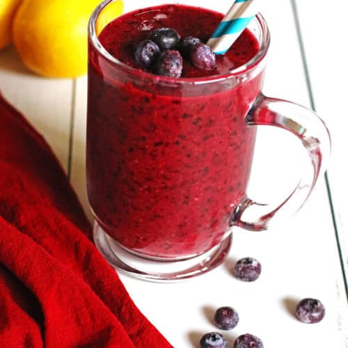 Smoothie made with blueberries and lemon in a clear mug sitting on a white table.