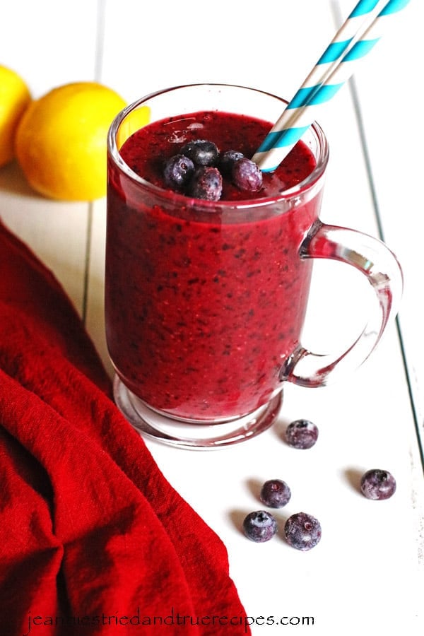 Smoothie made with blueberries and lemon in a clear mug sitting on a white table.