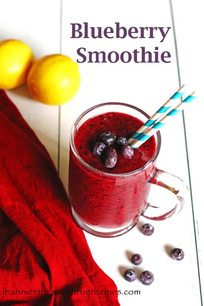 Blueberry Smoothie in a clear mug with a few frozen berries on top. Lemons and blueberries are on the table near the smoothie.