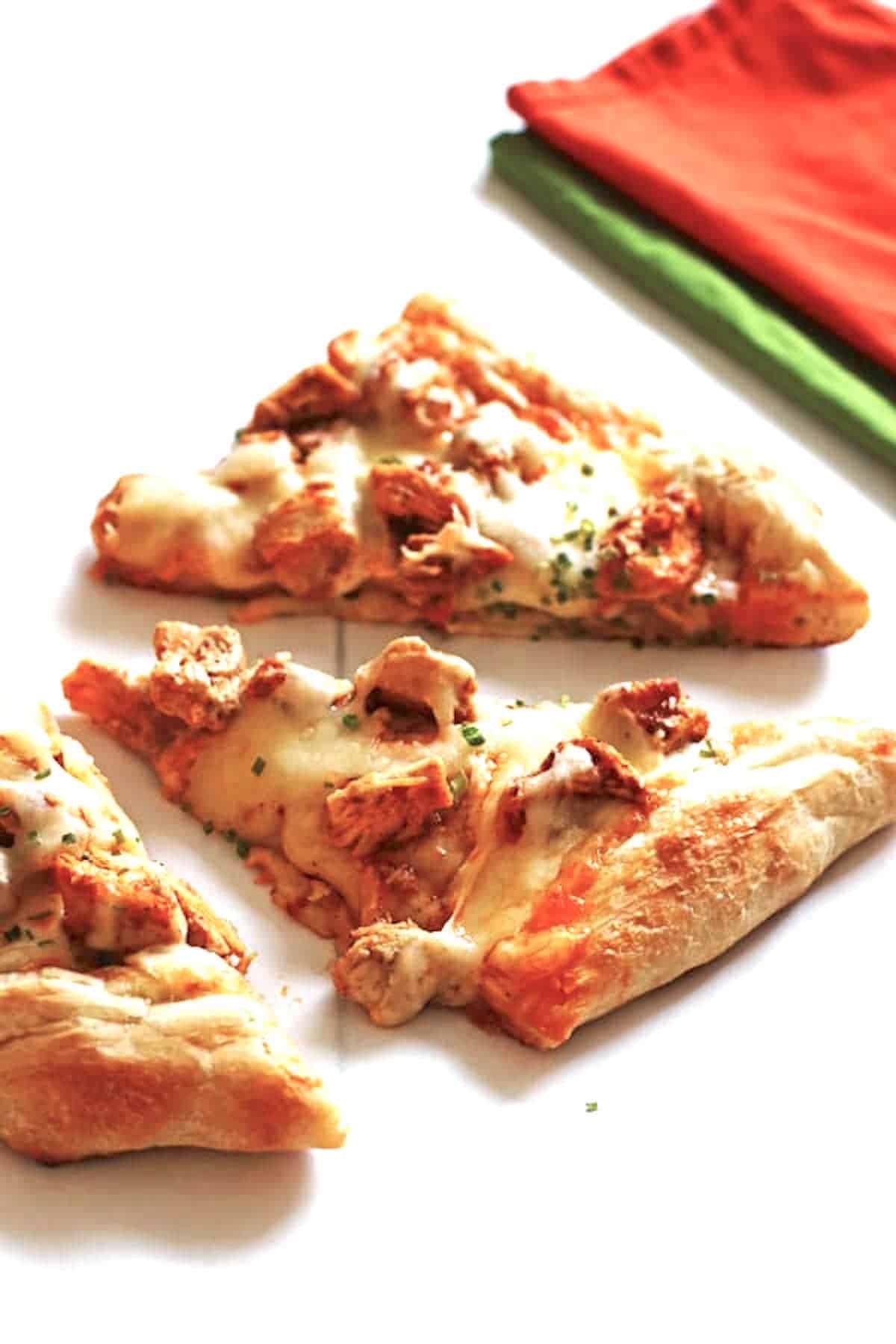 Pizza slices covered with cheese, chicken and chicken wing sauce on a white table.