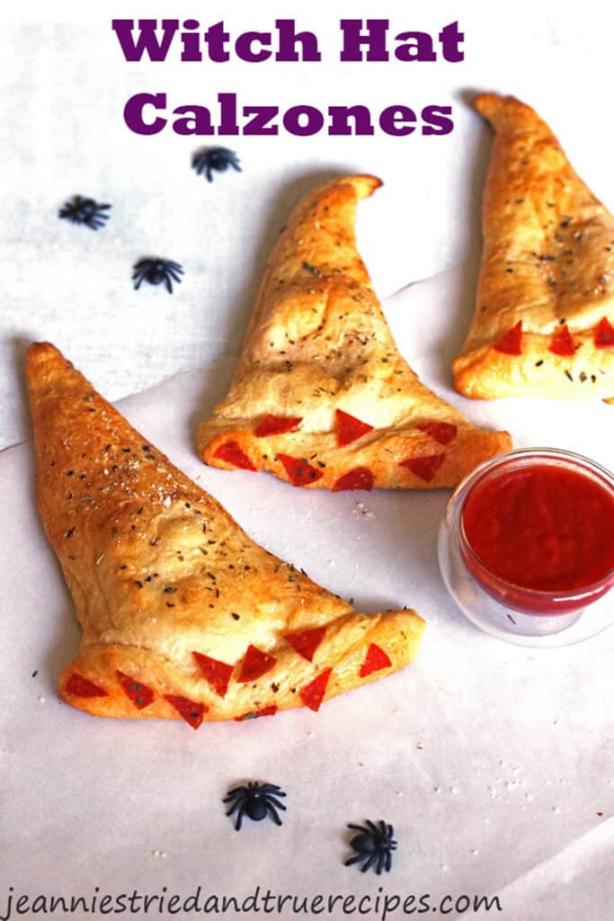 Calzones shaped as witch hats. They are placed on a white table with a small bowl of marinara sauce. There are also plastic black spiders as decor on the table.