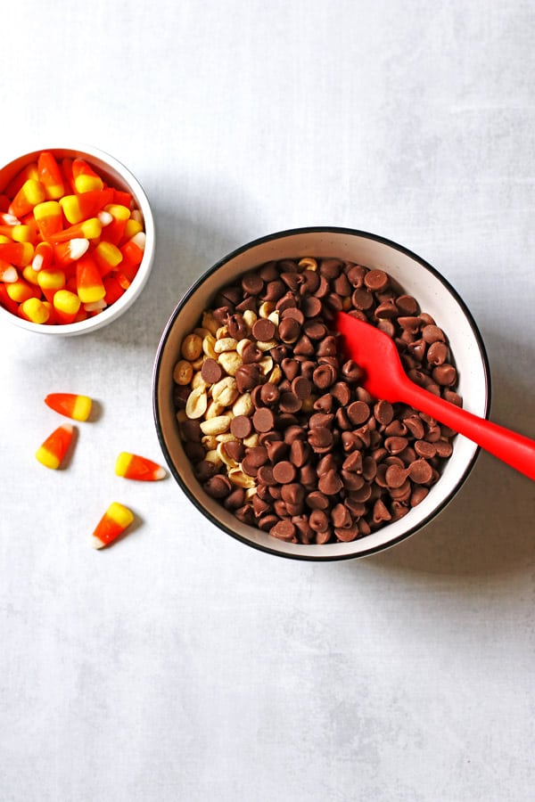 Chocolate chips and peanuts in a large white bowl and candy corn in a measuring cup next to the bowl.