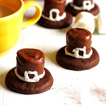 Cookies that are decorated as Pilgrim Hats sitting on a white table with a glass of milk