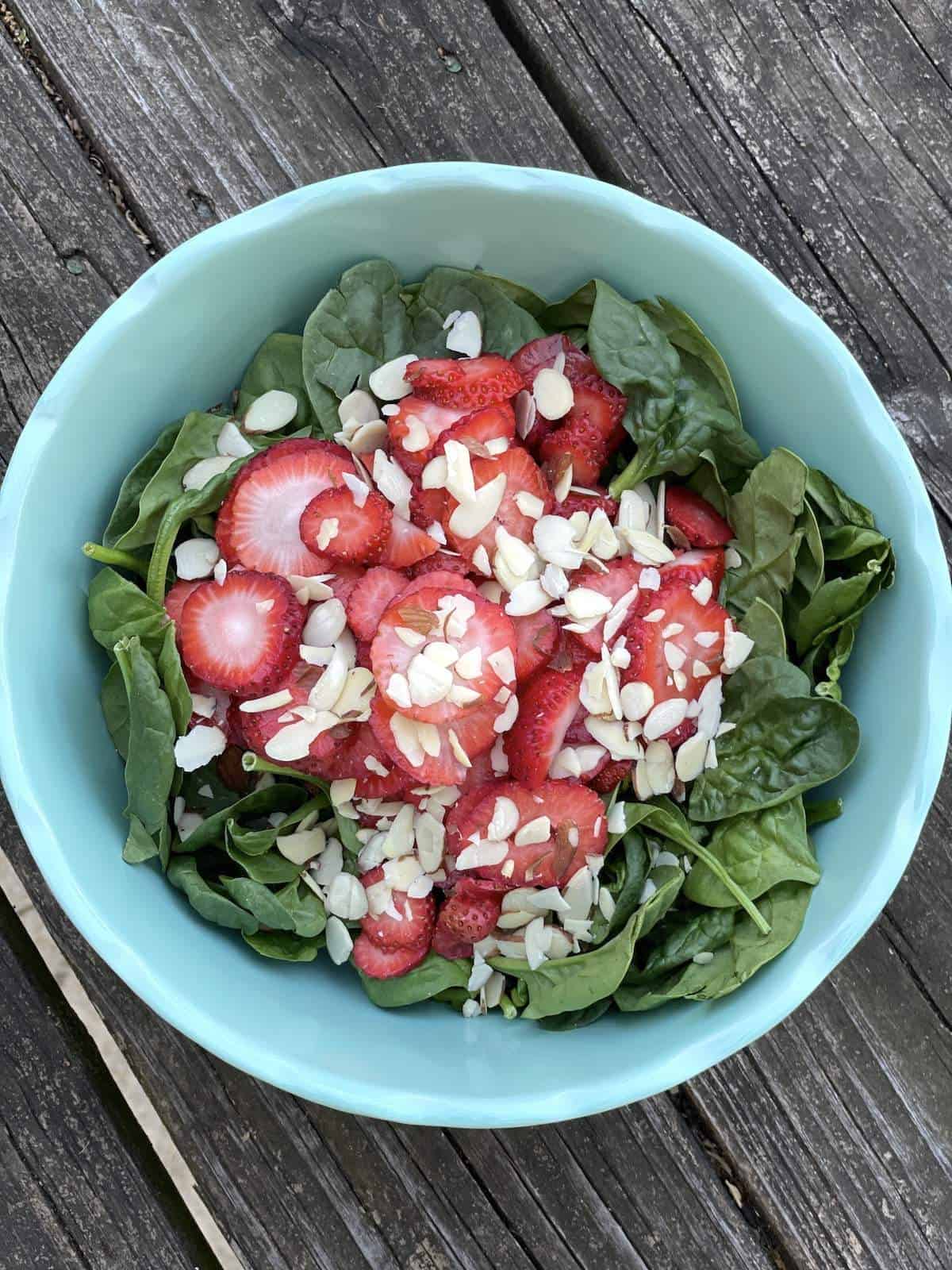 A bowl of spinach with strawberries and almonds on top for strawberry spinach salad.