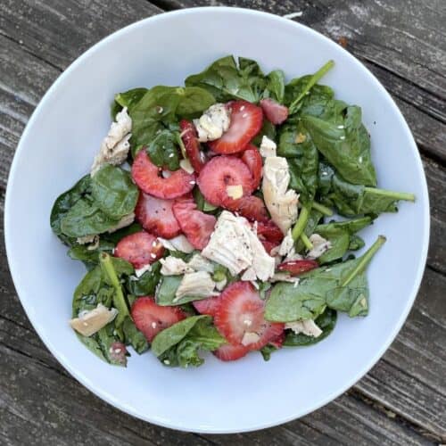 A white bowl of strawberry and spinach salad with almonds and chicken, tossed with sesame poppyseed dressing.
