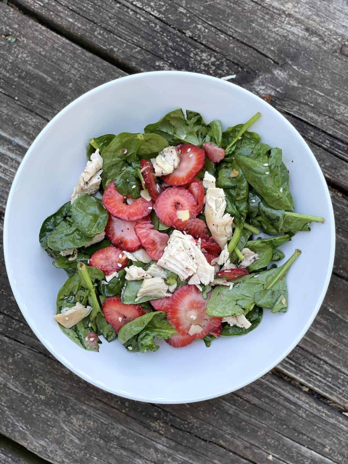 A white bowl of strawberry and spinach salad with almonds and chicken, tossed with sesame poppyseed dressing.