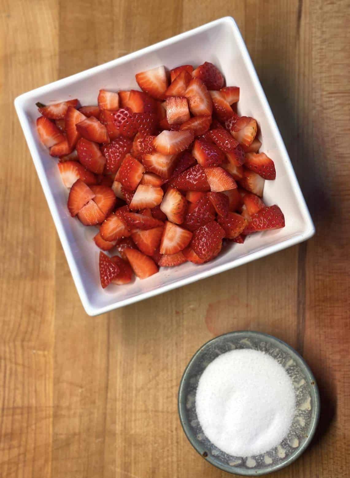 A bowl of strawberries and a bowl of sugar before being mixed together.