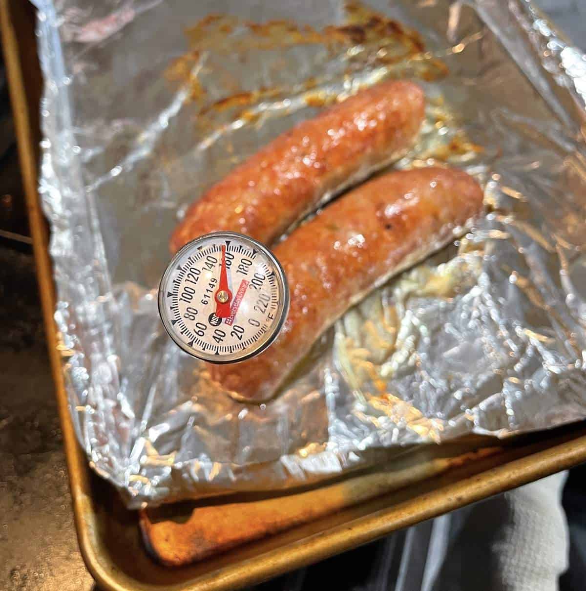 Two oven baked Italian sausages on a foil lined baking sheet with the temperature being taken.