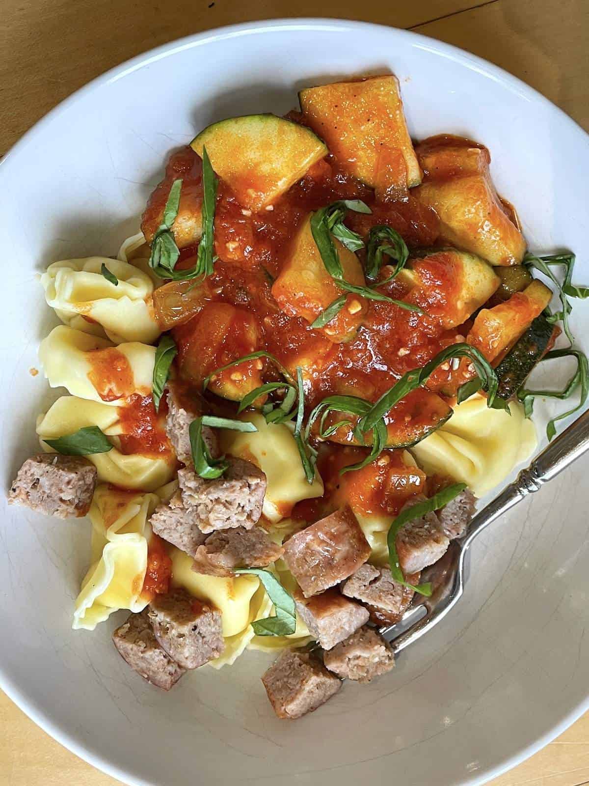 A bowl of cheese tortellini with oven cooked Italian sausahe, sauteed zucchini, homemade tomato sauce, & fresh basil.