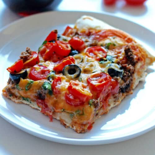 A square slice of taco pizza topped with cheese, black olives, and tomatoes.