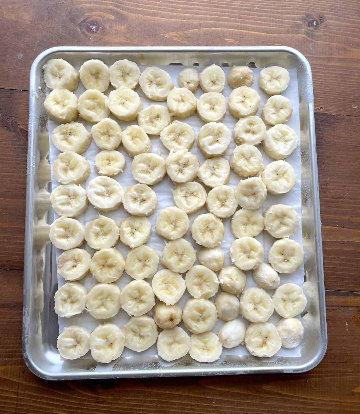 Bananas slices frozen in a single layer on a parchment paper lined baking sheet.