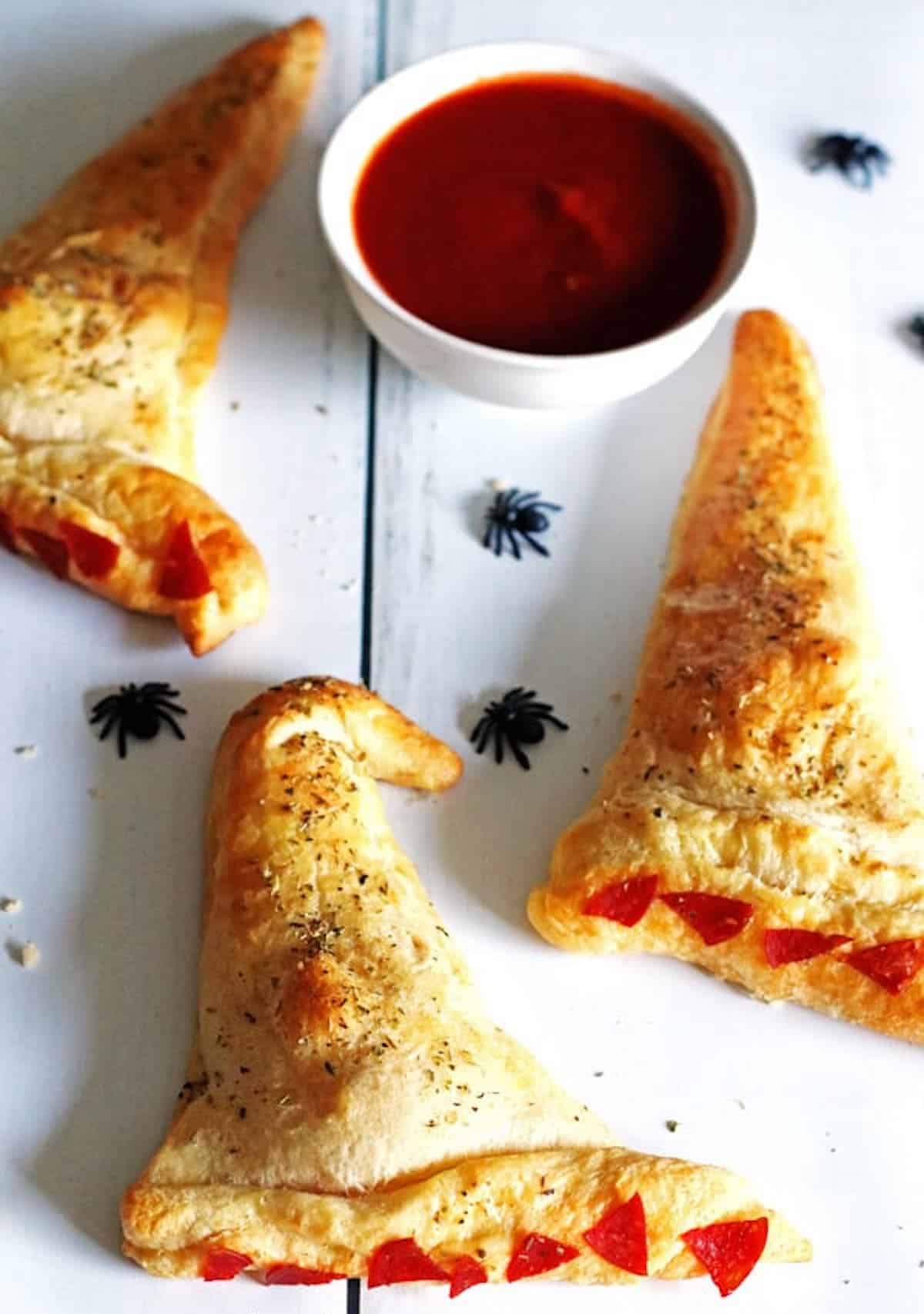 Three lightly baked witch hat calzones with plastic spiders and marinara sauce.