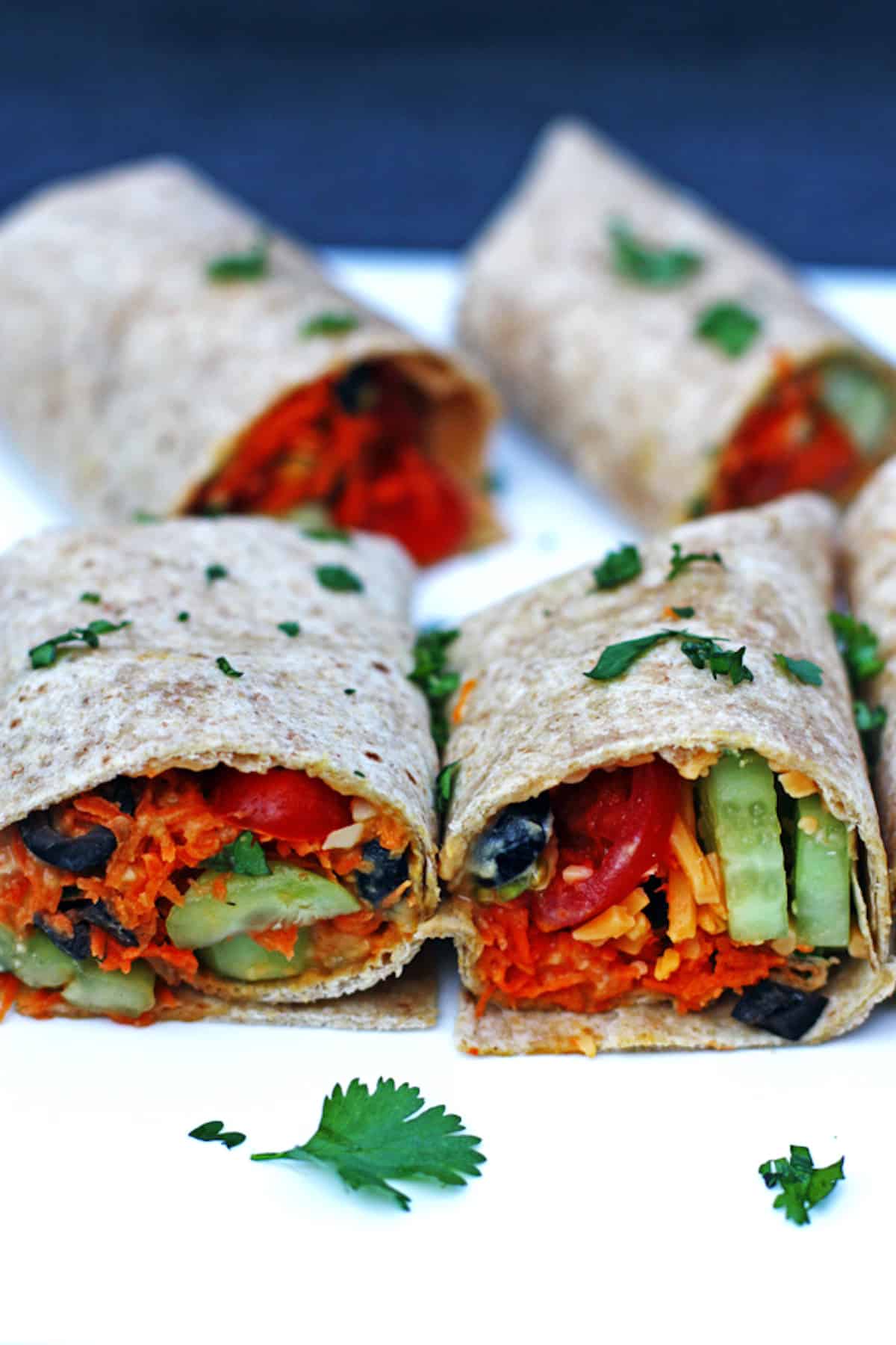 Four whole wheat vegetable wraps with tomatoes, hummus, carrots, cilantro, olives, and cucumber.