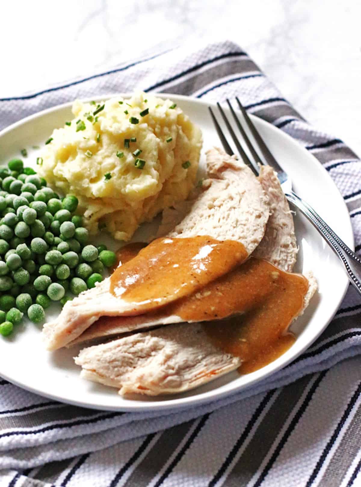 A plate of crockpot turkey breast with mashed potatoes and peas, smothered in gravy.