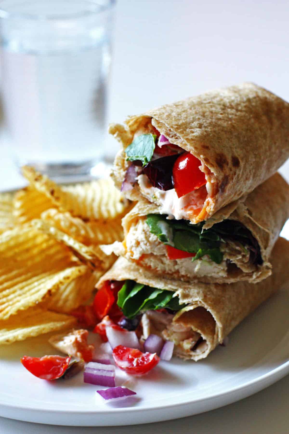 A stack of three chicken wraps on w plate with tomatoes, red onion, and chips.
