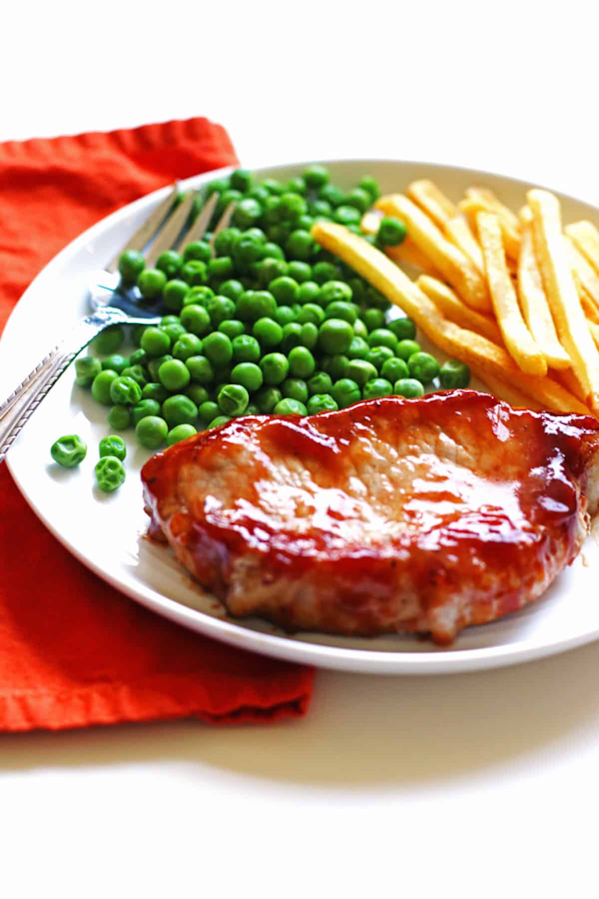 A white plate with a barbecue pork chop, peas, and french fries.
