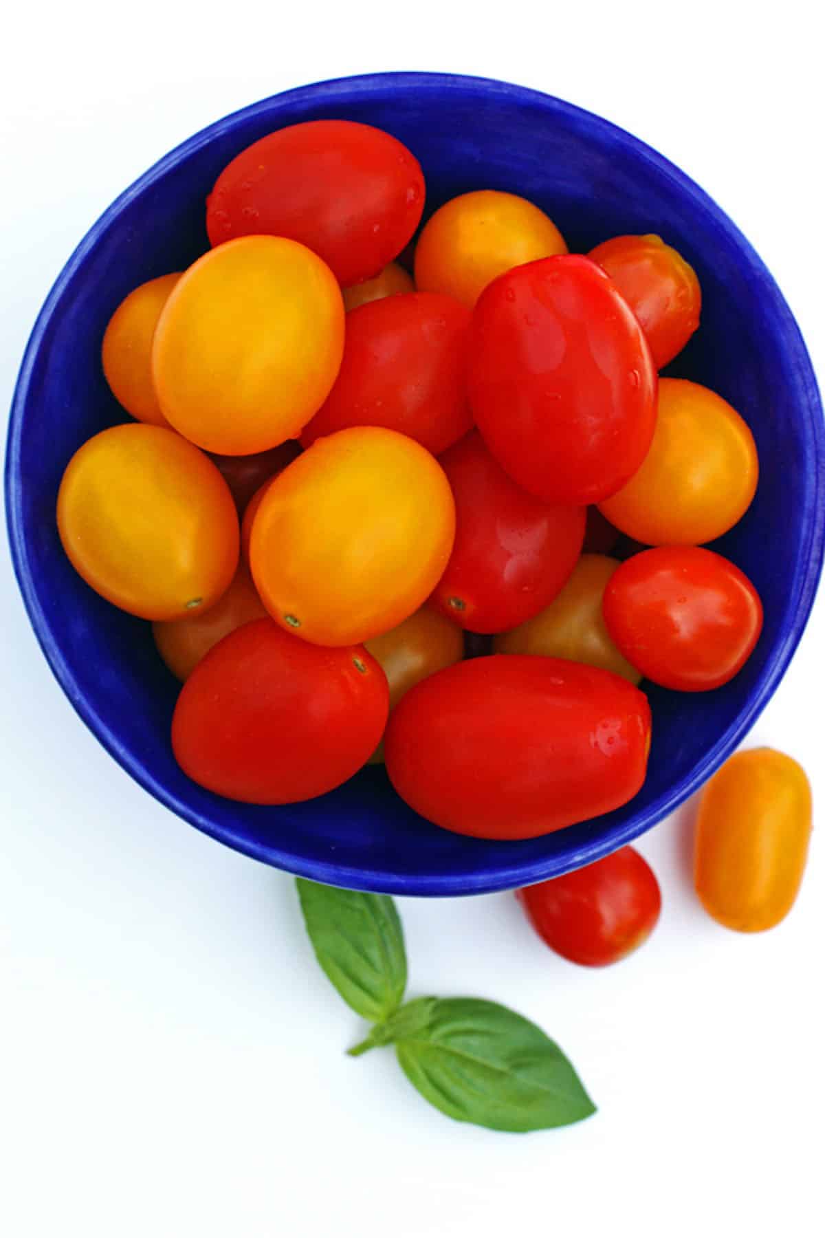 A blue bowl of cherry tomatoes with basil on the side.