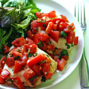 A plate of bruschetta chicken with mozzarella, tomatoes, and basil on top and a side salad.