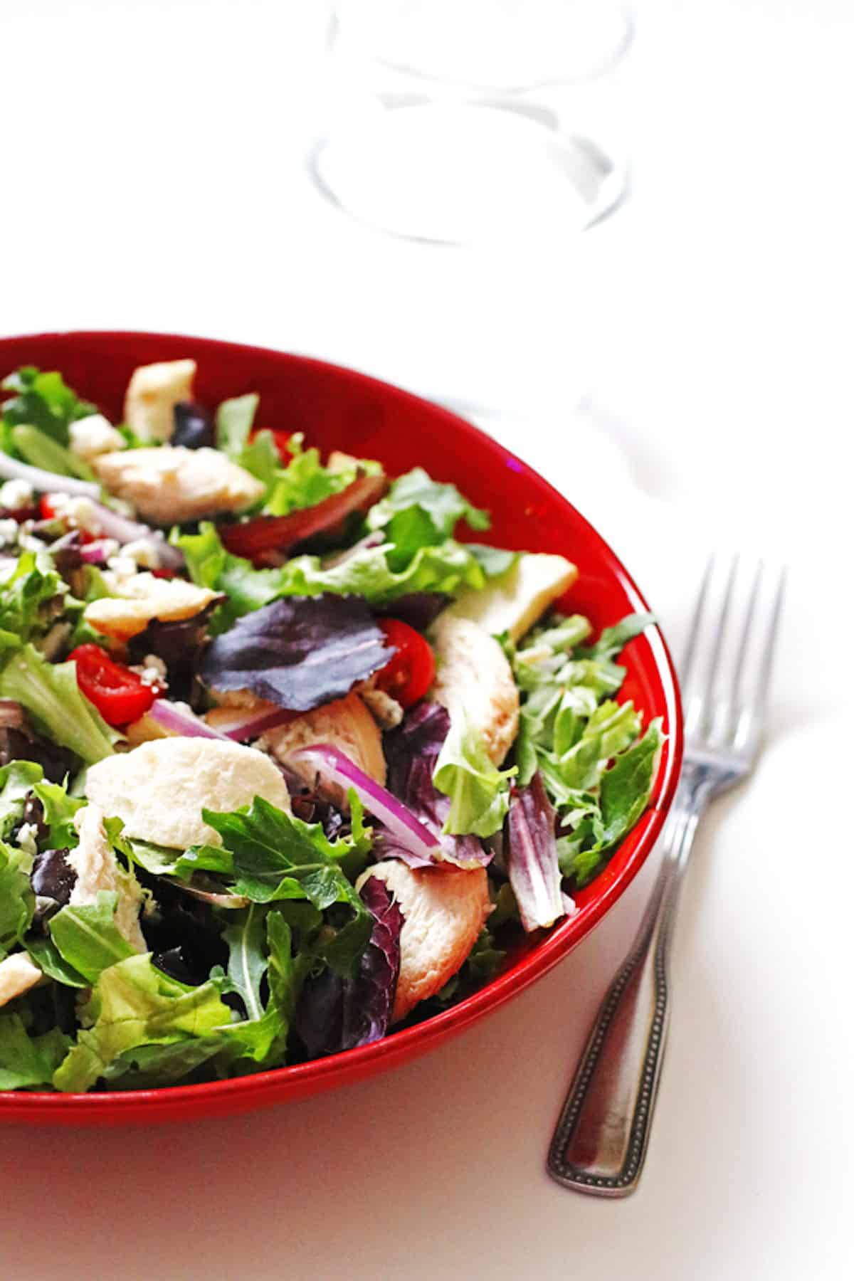A red bowl of panera fuji apple chicken salad with red leaf lettuce and greens, red onions, tomatoes, chicken, apple chips, gorgonzola, and white balsamic dressing.