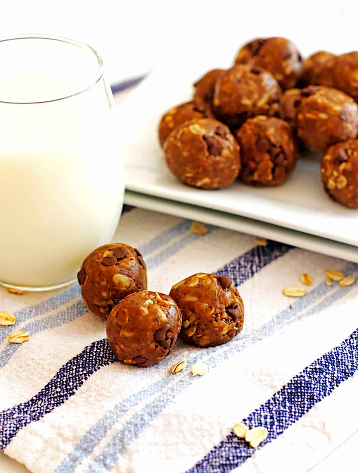 A plate of peanut butter energy bites on a white plate being picked up by a hand with a glass of milk and three in the front.
