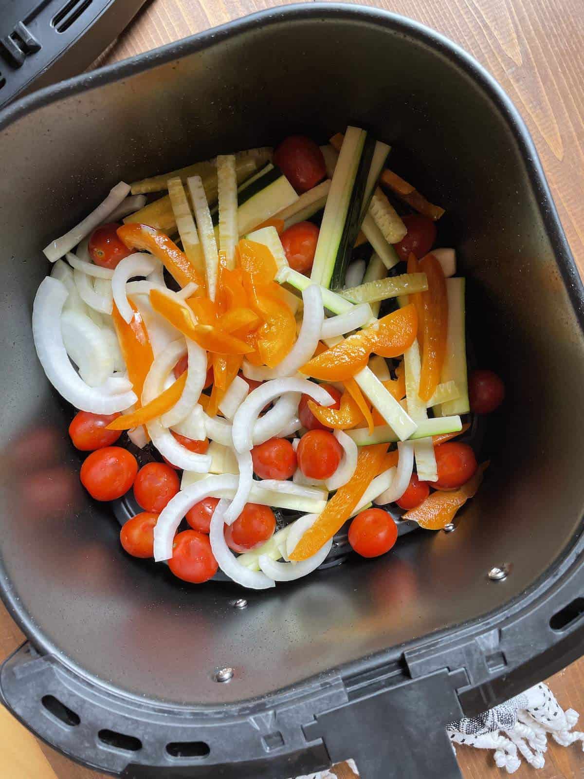 Peppers, onions, zucchini, and tomatoes cooking in an air fryer basket.