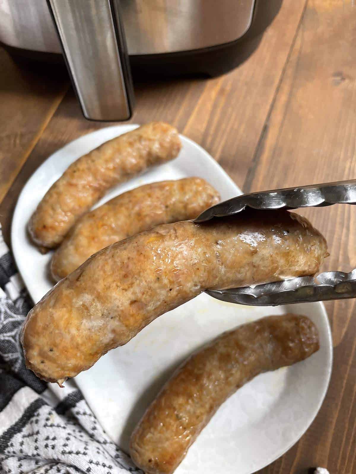 A pair of tongs holding an air fried Italian sausage over a white plate.