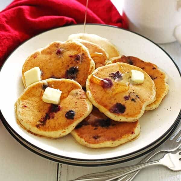 Pancakes made with blueberries on a white plate with sliced butter on top of them and syrup being poured over them