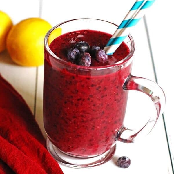 Smoothie made with blueberries and lemon in a clear mug sitting on a white table.