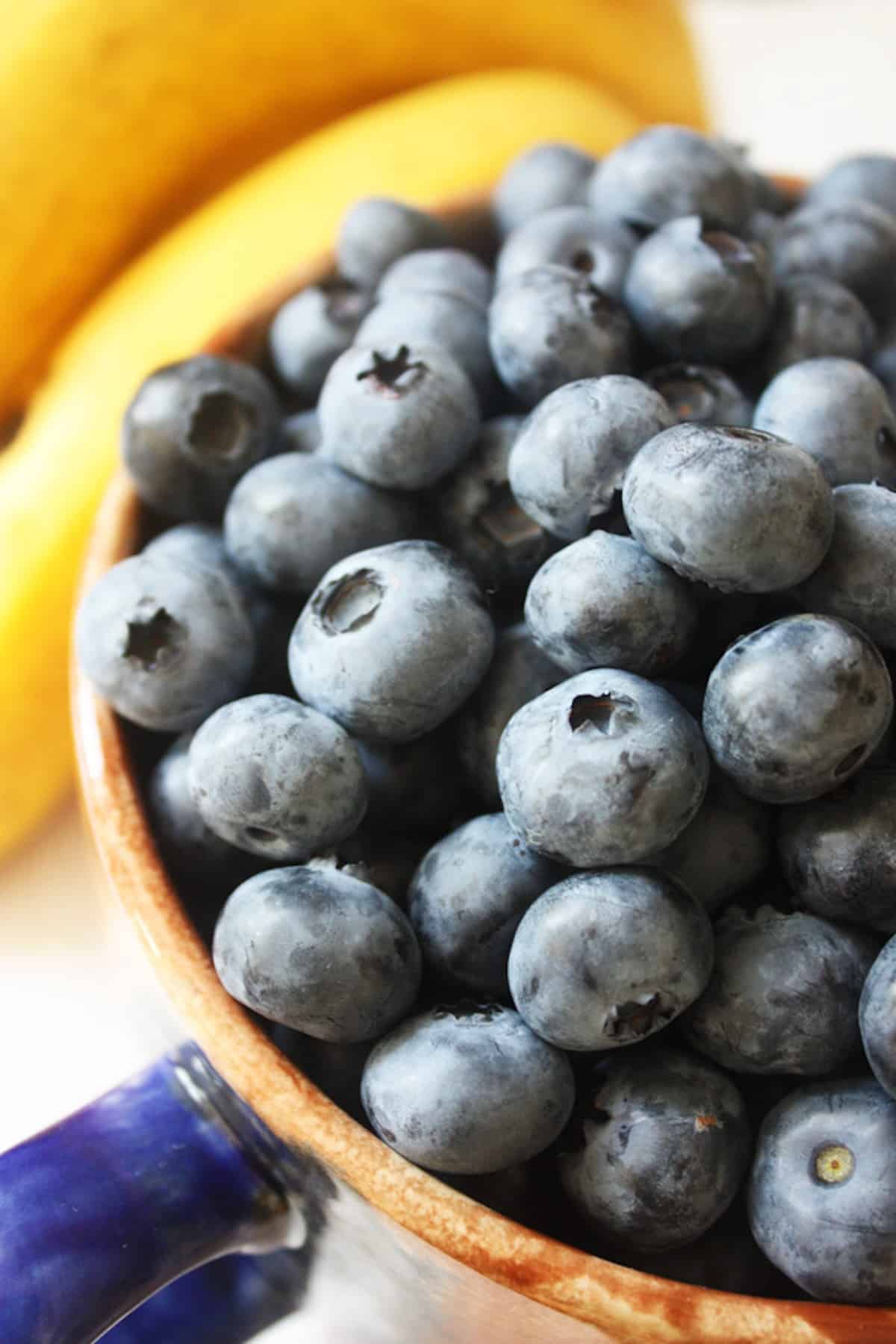 A bowl of blueberries with bananas in the background