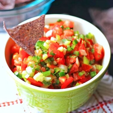 A green bowl of fresh salsa with cilantro, diced tomatoes, and peppers with a chip sticking out of it.